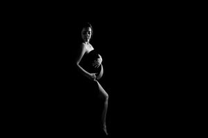 Artistic black and white maternity silhouette shows a pregnant figure dramatically posed against a dark background.