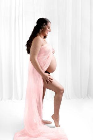 Graceful maternity silhouette against white curtains wearing flowing pink fabric in artistic pregnancy photoshoot.