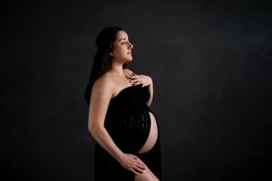 A dramatic maternity portrait against a dark background shows an expectant mother draped in black fabric.