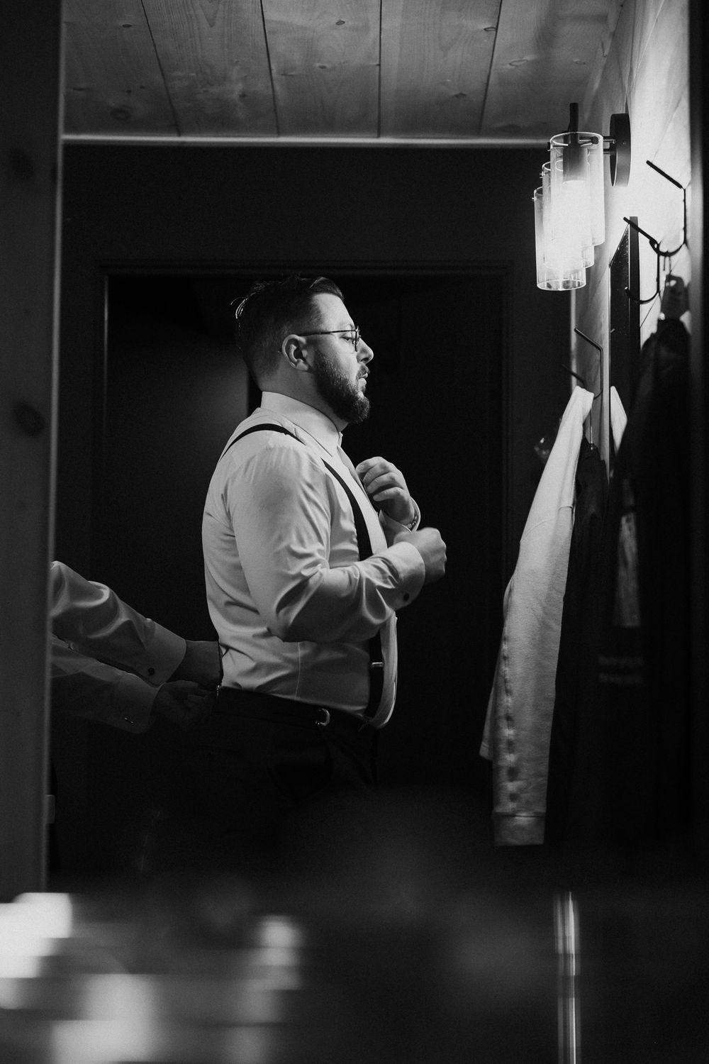 A black and white photograph shows someone in suspenders and button-up shirt adjusting their tie in dim lighting.