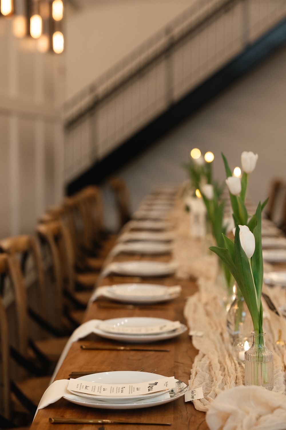 Elegant table setting with white tulips in glass vases and place settings along a rustic wooden dining table with vintage chairs.