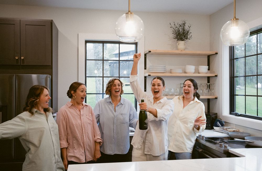 A group of coworkers laughing together in a bright modern kitchen with pendant lights and floating shelves.