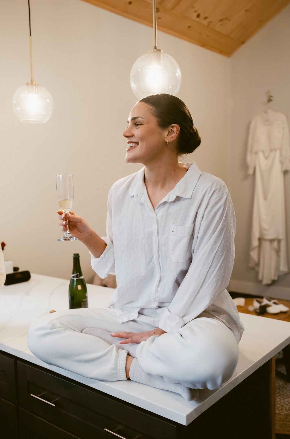Someone wearing white pajamas sits cross-legged on bed holding champagne glass with pendant lights and robe in background.