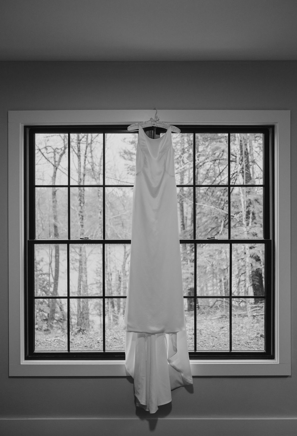 A white wedding dress hangs elegantly in front of a large windowpane overlooking a winter forest scene.
