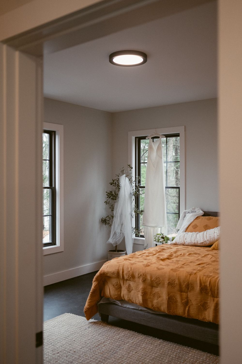 Modern bedroom with golden brown bedding, white walls, and an elegant hanging light fixture near tall windows.