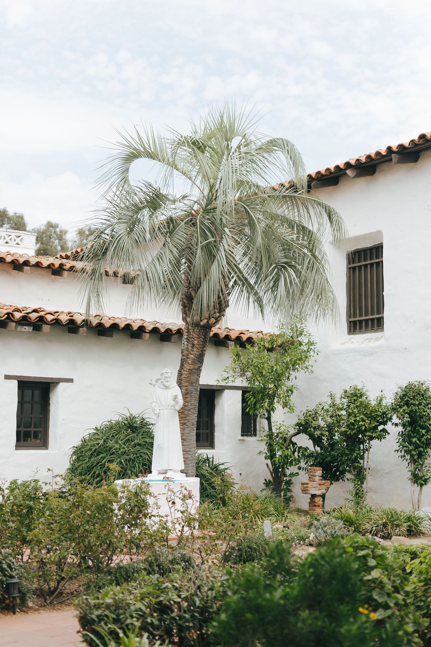 Spanish colonial style white building with palm trees and lush garden landscaping along terracotta tile walkway.