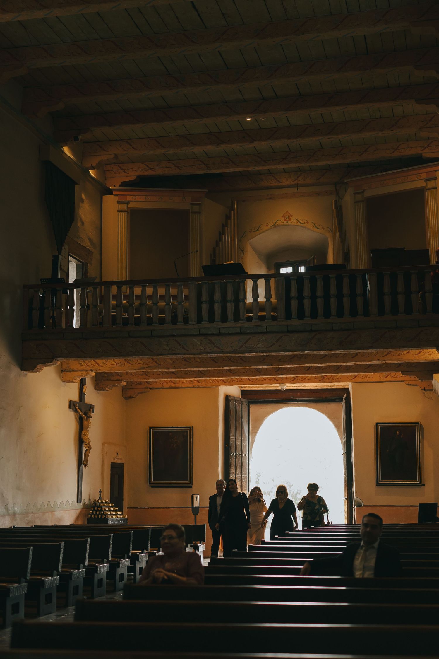 Silhouettes of people walking through a sunlit doorway in a historic church with balcony.