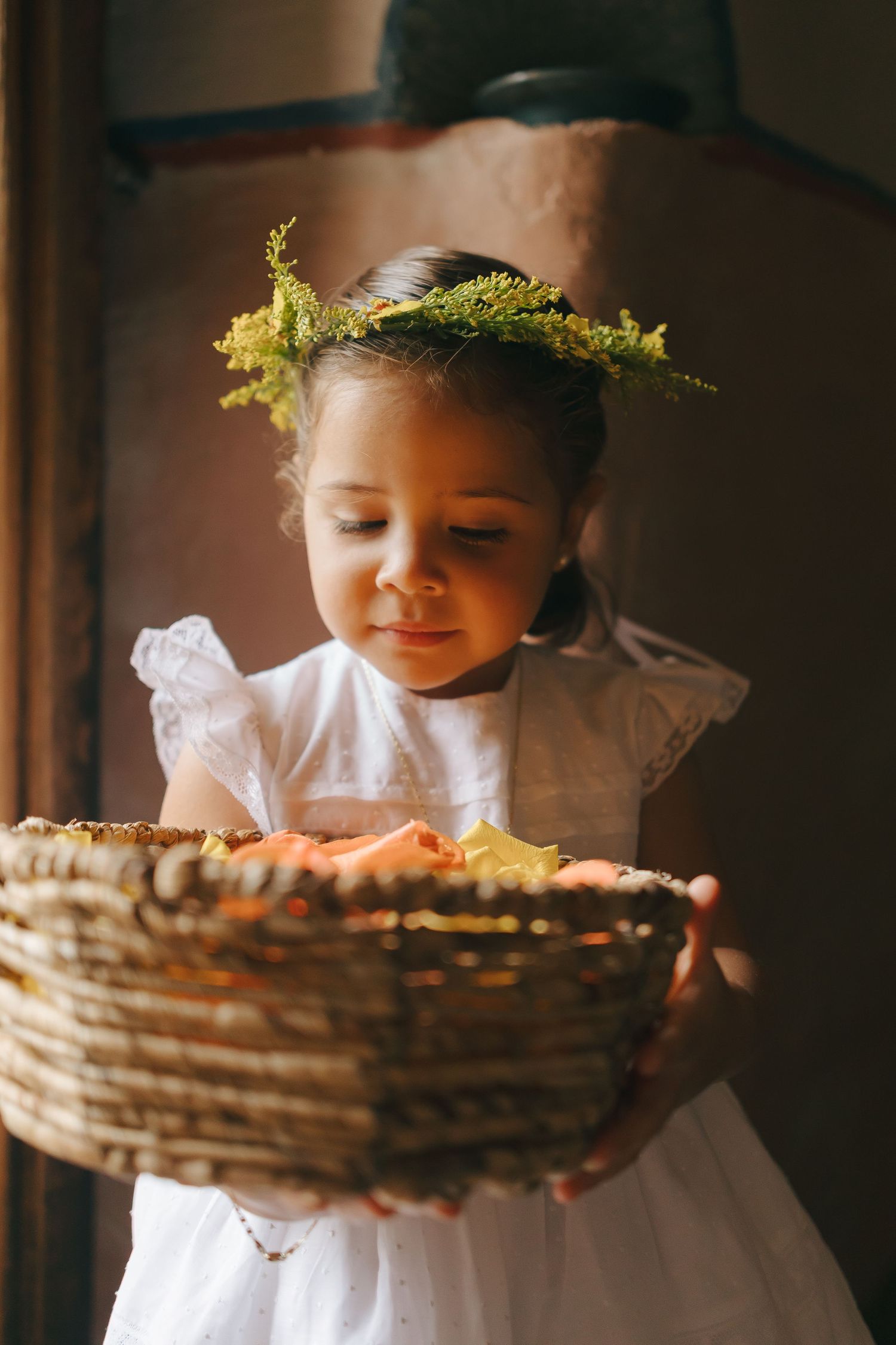 A young attendant in white dress wearing floral crown holds woven basket.