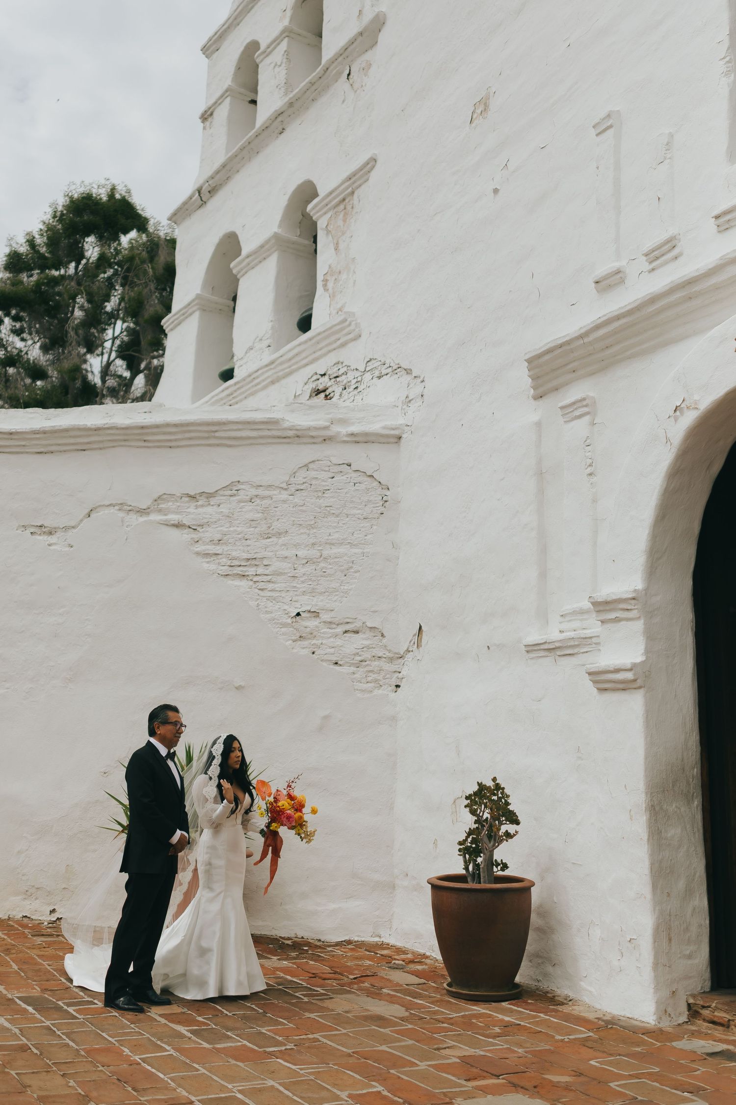 A wedding couple stands against a white Spanish colonial mission church wall with arched bell towers.