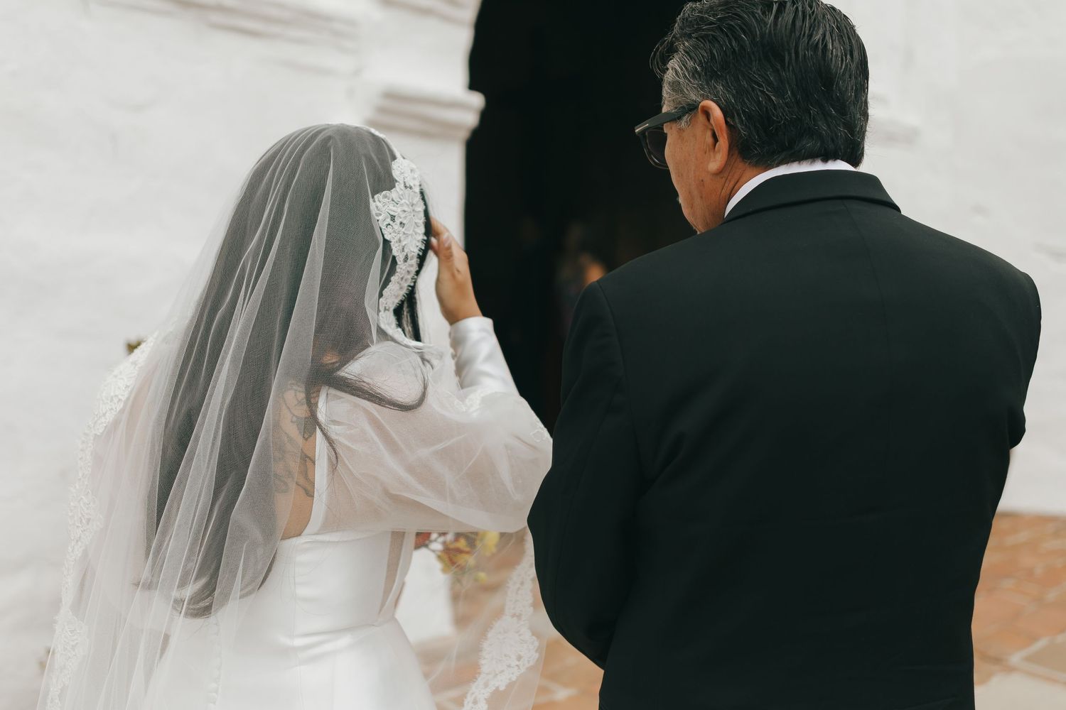 A bride in a white wedding dress touches her veil while a person in a dark suit stands nearby during the ceremony.