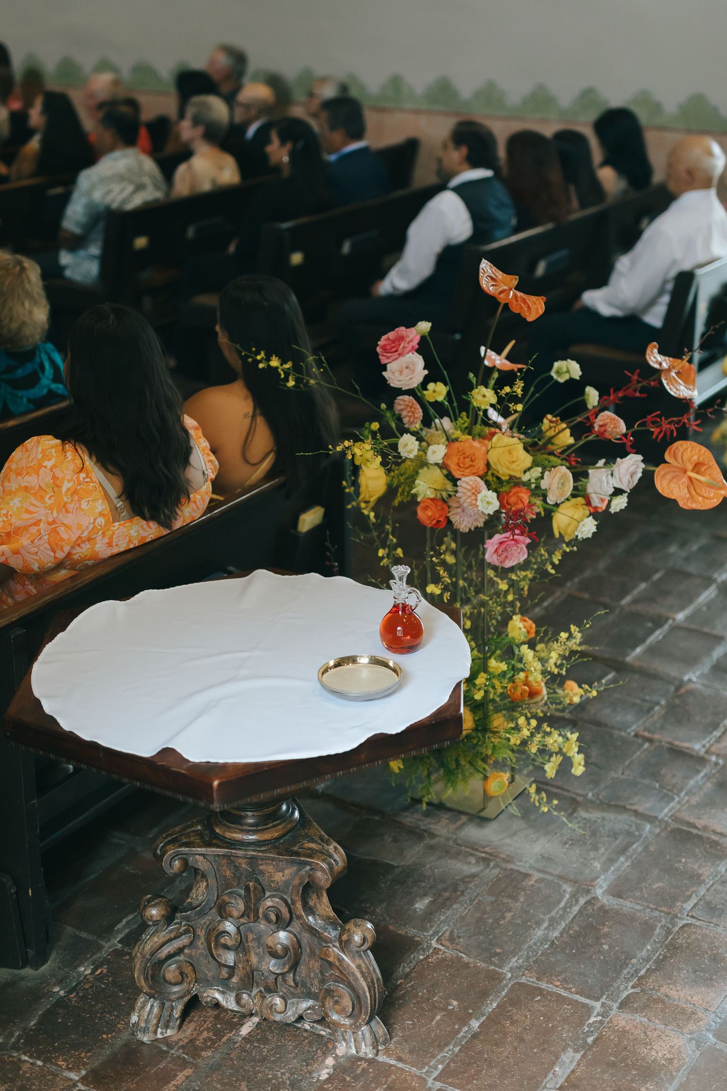 Elegant floral centerpiece and white cake plate set against a backdrop of wedding reception guests in formal attire.