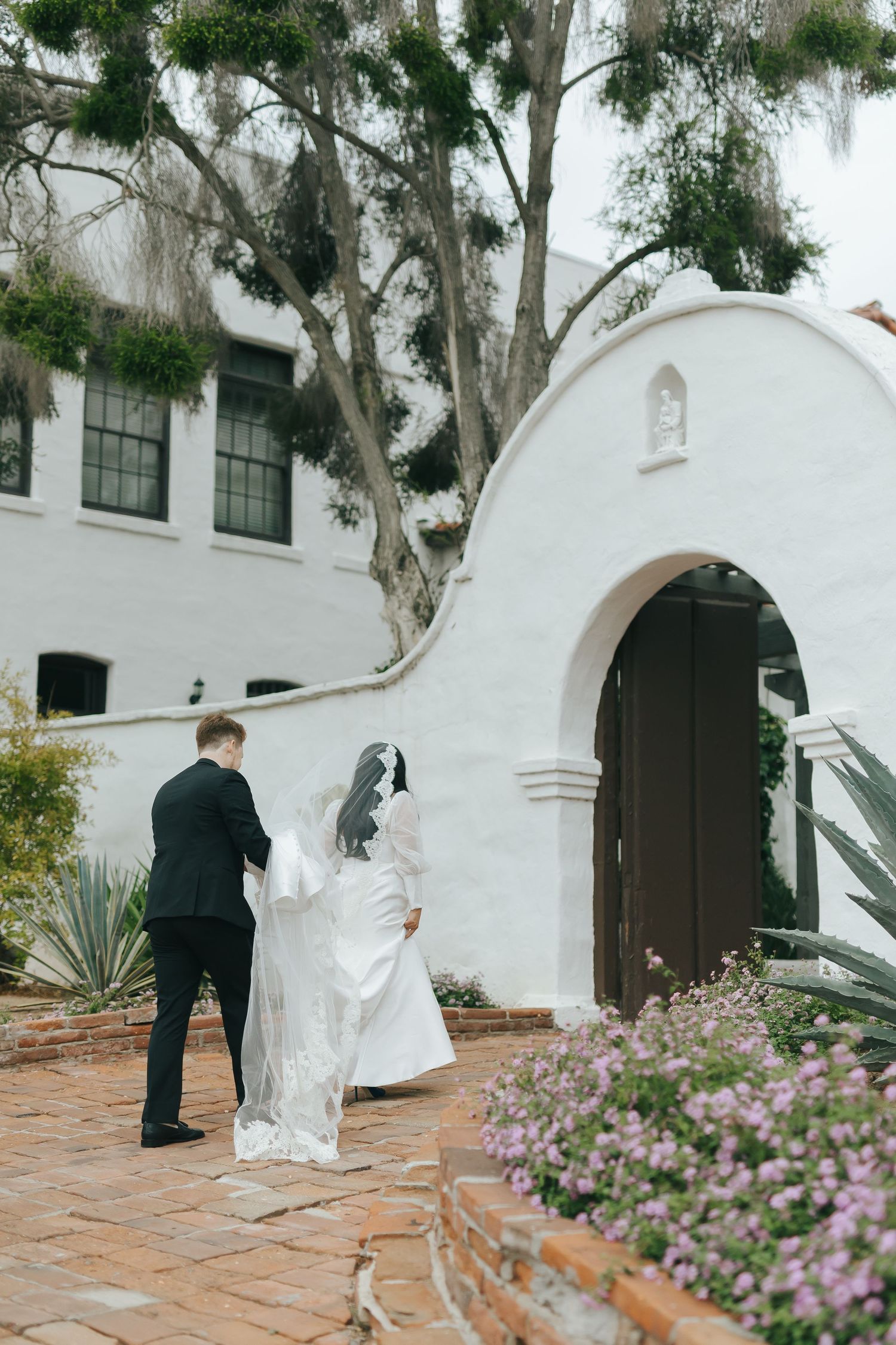 A couple entering through a white Spanish-style arch doorway with flowering plants and eucalyptus trees.