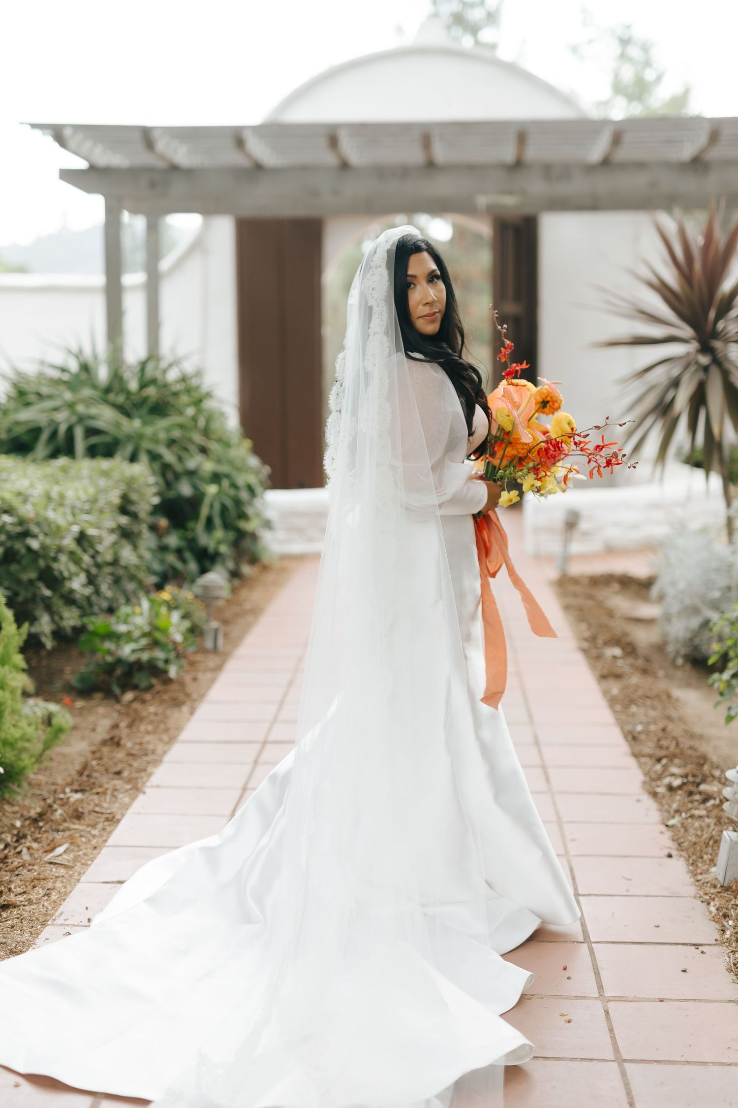 Bride in a flowing white dress holds vibrant orange and coral bouquet in a garden setting.
