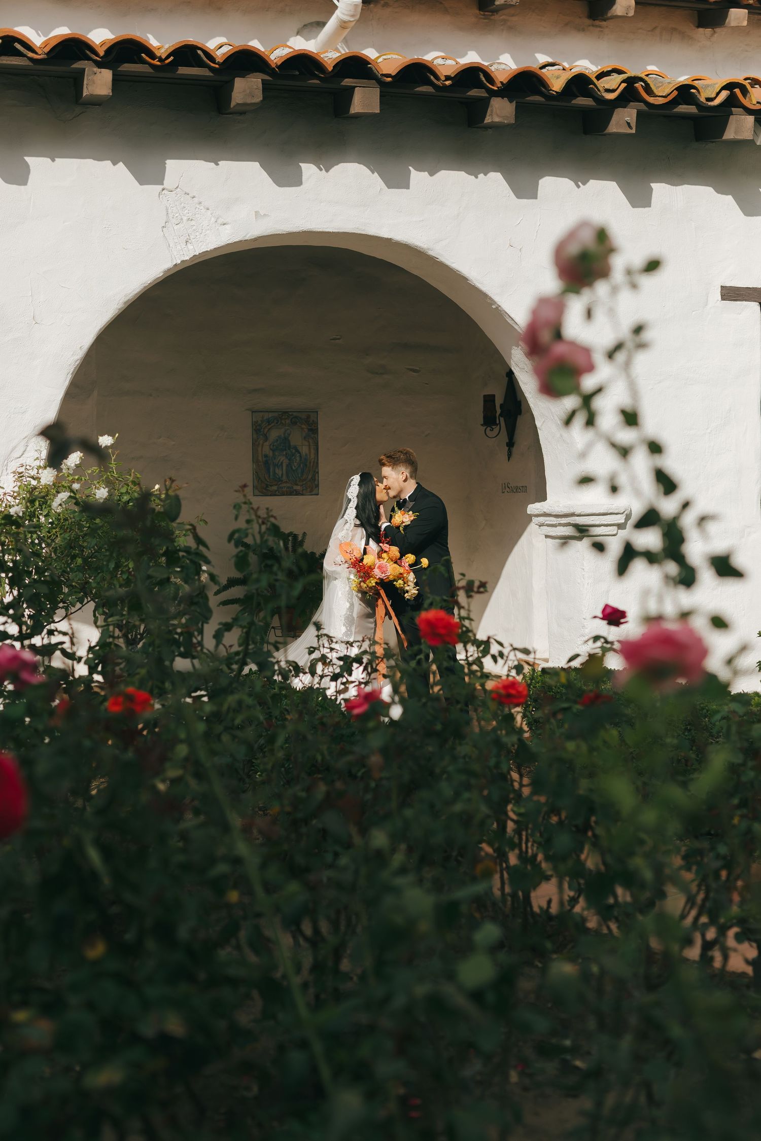 A romantic wedding scene in a Spanish-style courtyard with white walls and blooming rose garden.