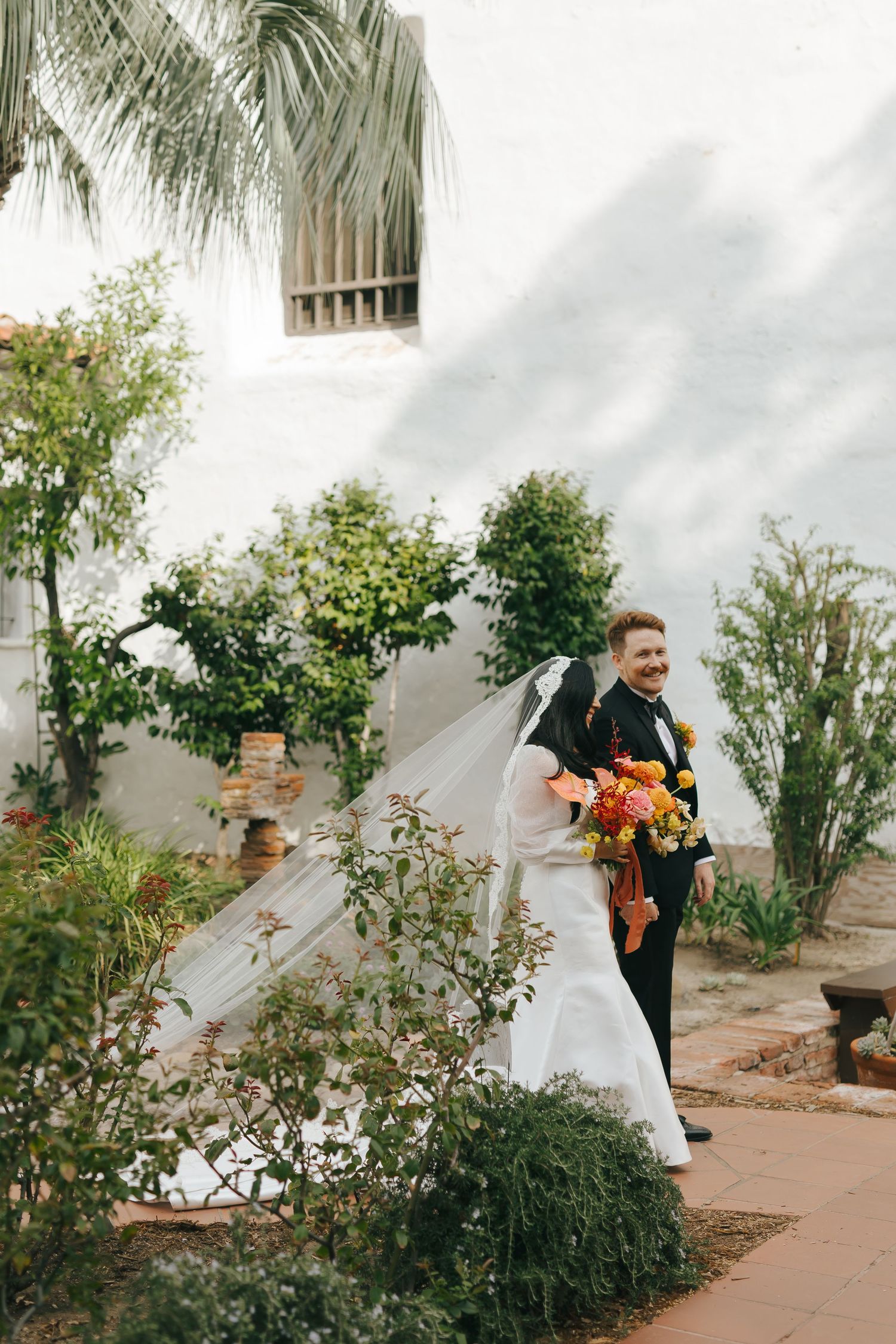 Newlyweds share a joyful moment in a Mediterranean garden with palm trees and white stucco walls.