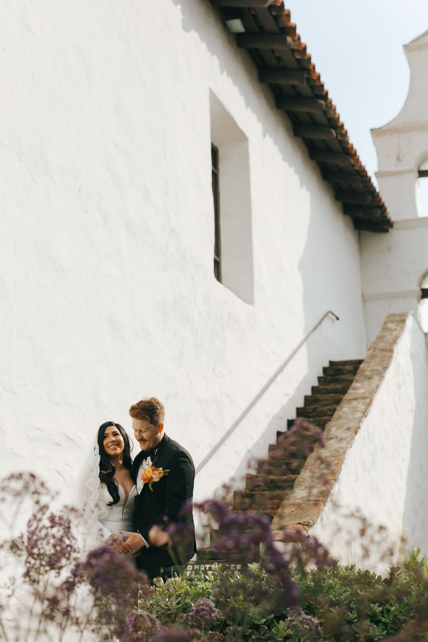 A romantic wedding moment captured against white mission-style architecture and flowering shrubs.