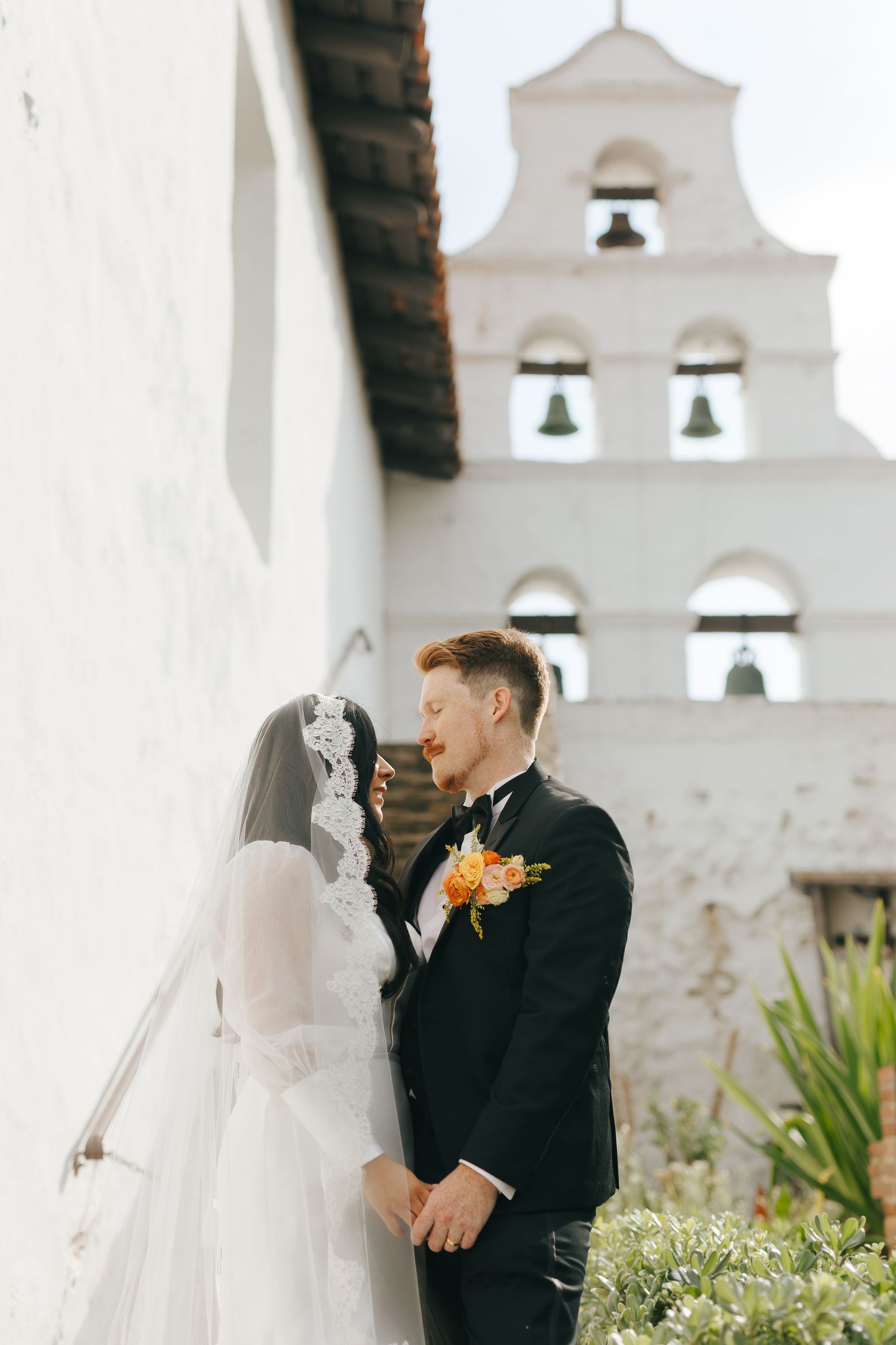 Newlyweds share a romantic moment in front of a Spanish mission-style church with bell tower.