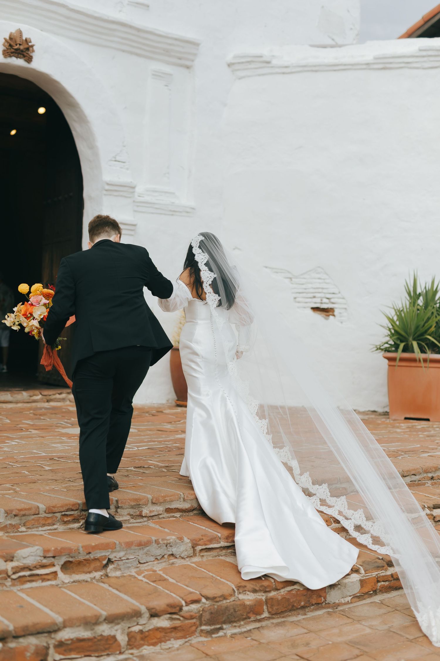A couple ascending brick steps at a historic white mission church during their wedding ceremony.