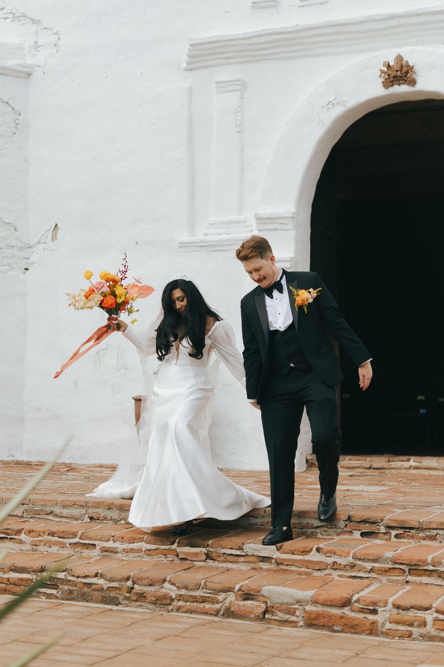 Wedding couple gracefully walking along historic white mission archways and terra cotta steps.