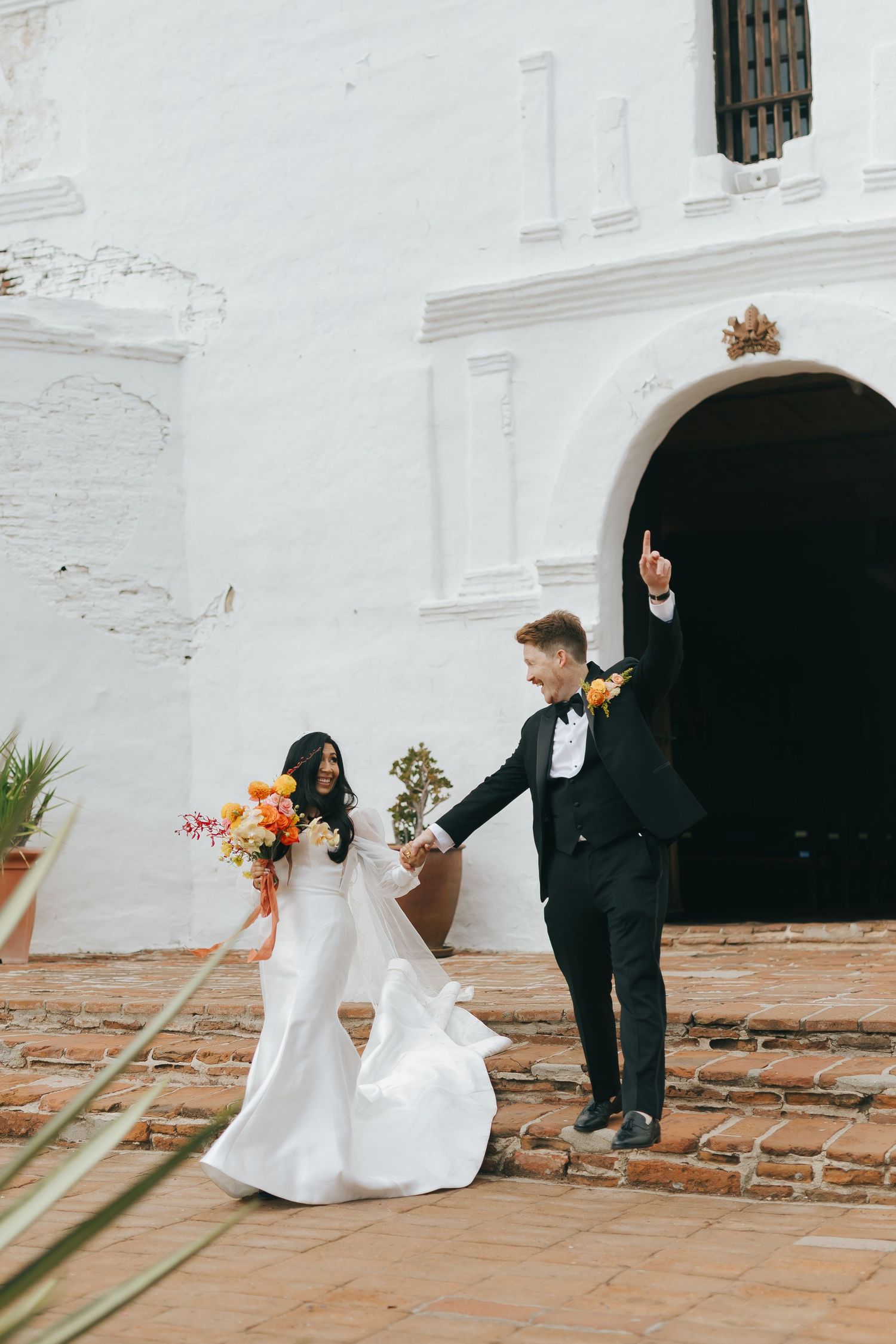 Bride and groom walk hand in hand on brick steps outside a white Spanish colonial church.
