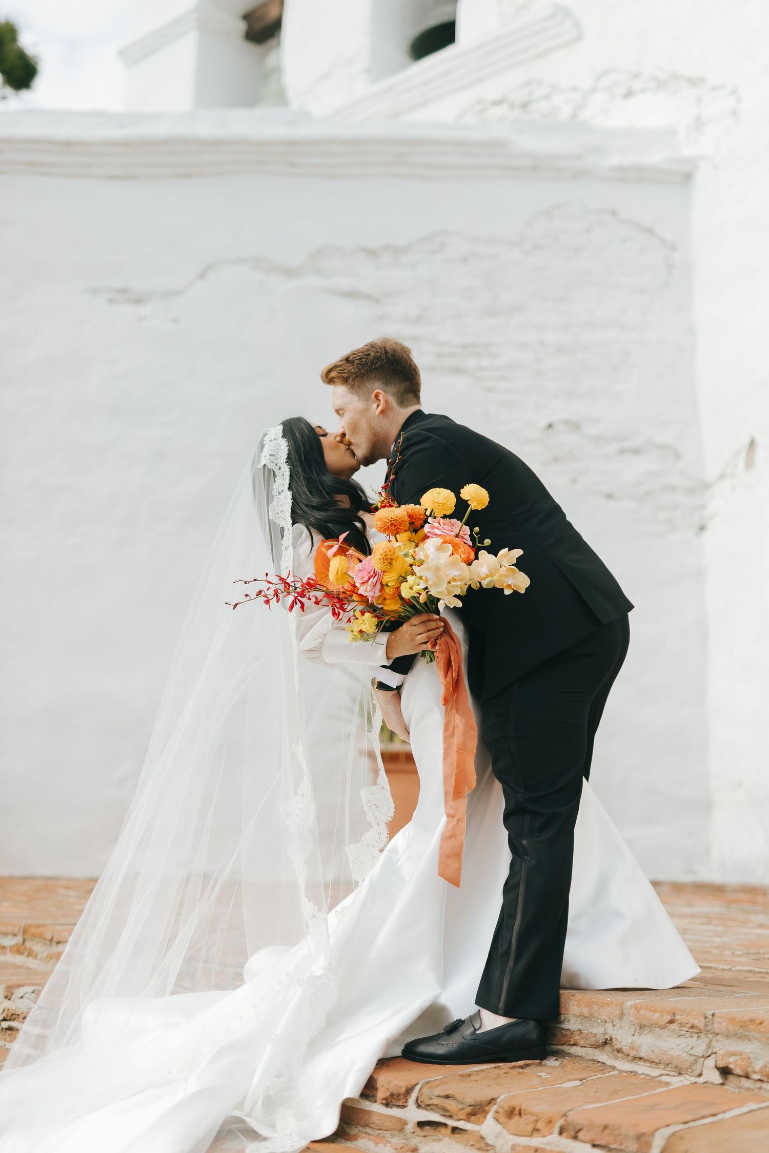A tender moment captured during a wedding ceremony against a white stucco wall with vibrant orange bouquet.