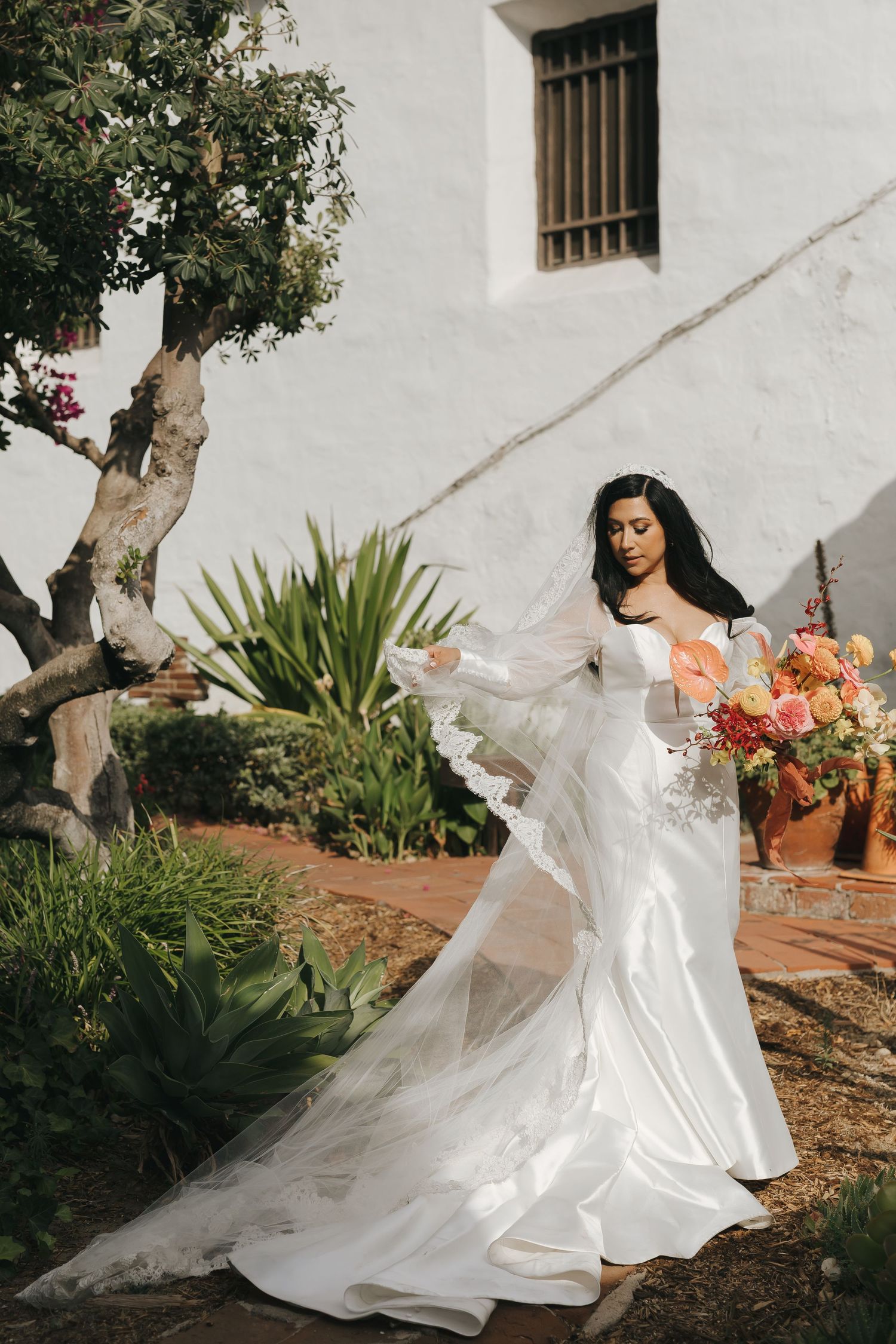 Elegant bridal portrait with flowing white gown and veil against Spanish mission style architecture.