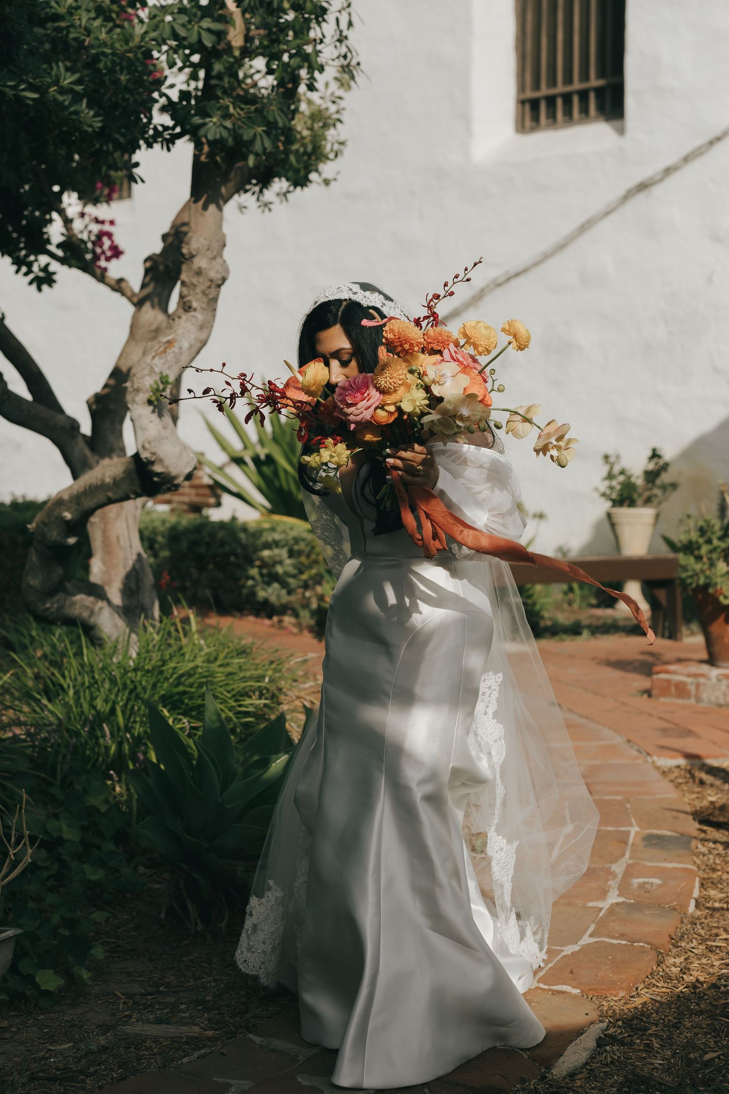 Flowing white wedding dress and colorful bouquet against garden pathway with Mediterranean architecture.
