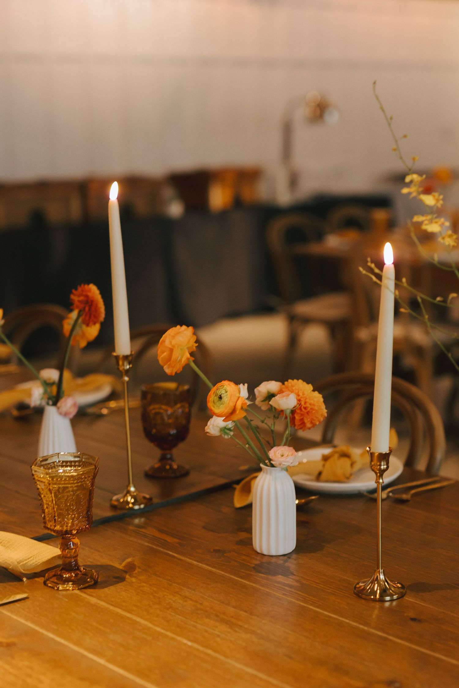 Romantic table setting with white candles, orange flowers, and golden glasses on wooden surface.