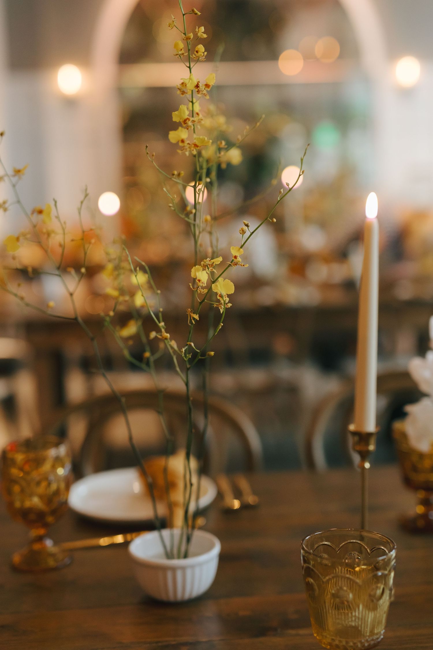Elegant wedding reception table with yellow flower stems in vase, candlesticks and amber glassware.