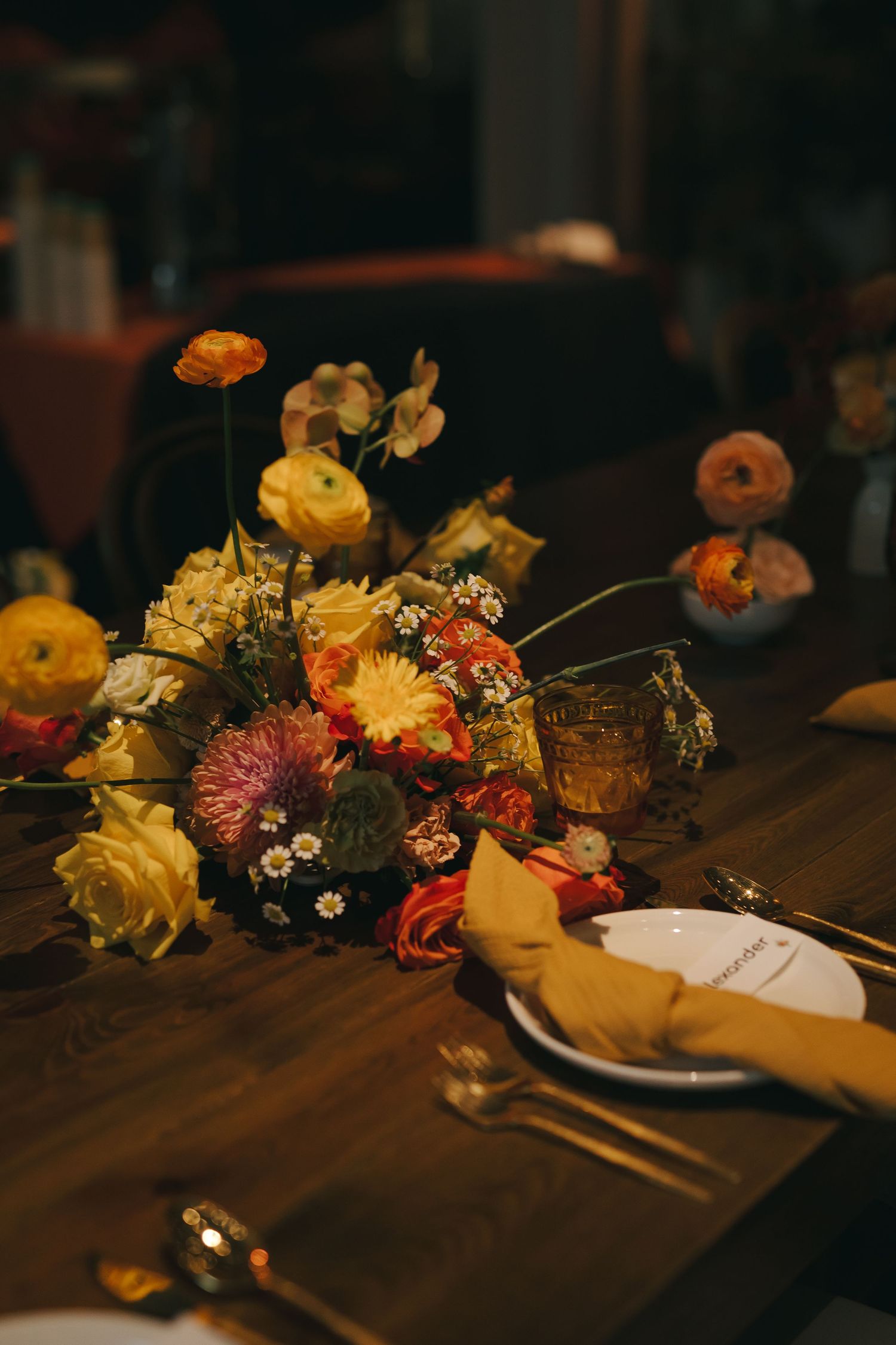 A romantic table setting with vibrant yellow and orange floral centerpiece on dark wooden surface.