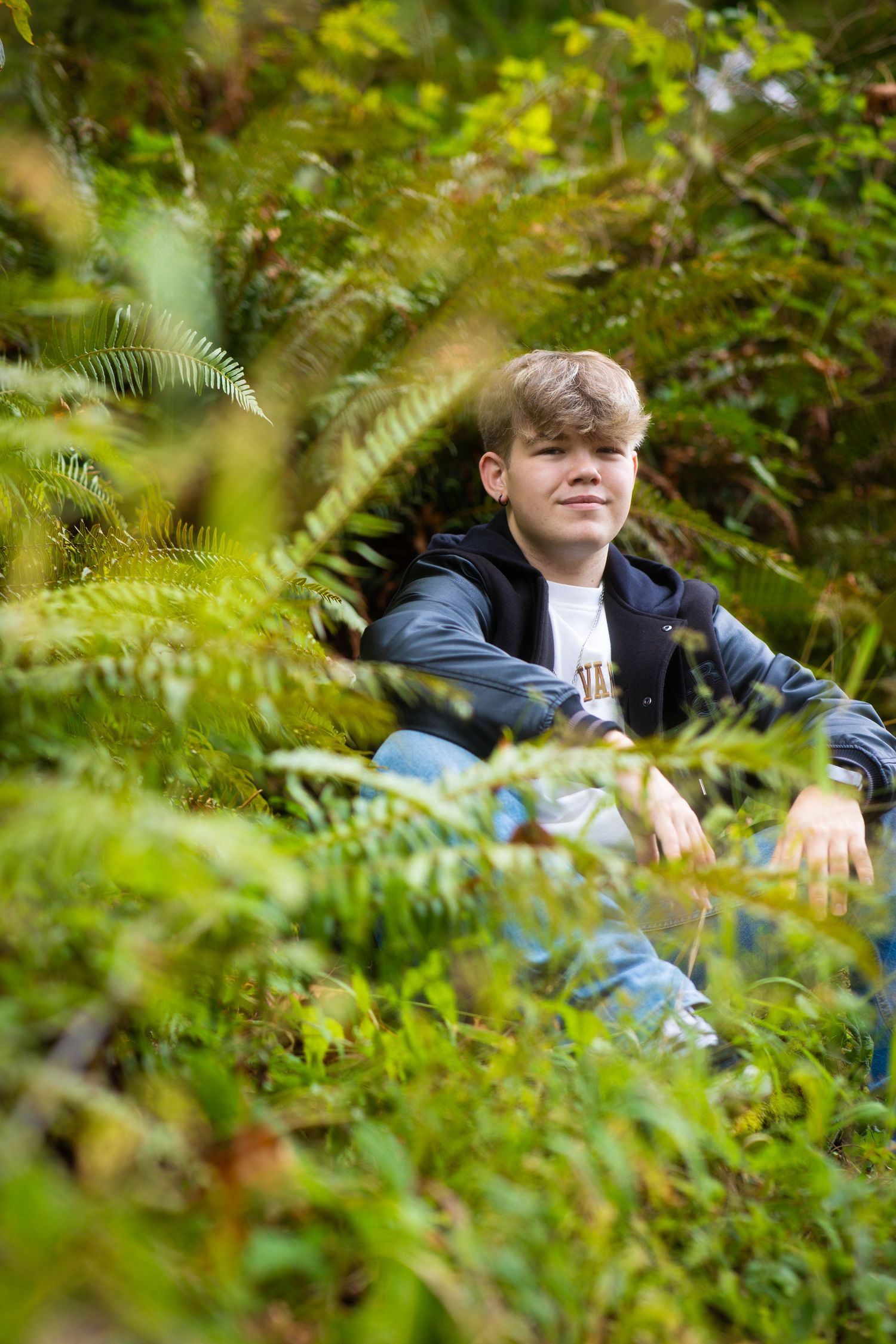 A person sits among vibrant green ferns and foliage wearing a navy blue jacket in a natural outdoor setting.
