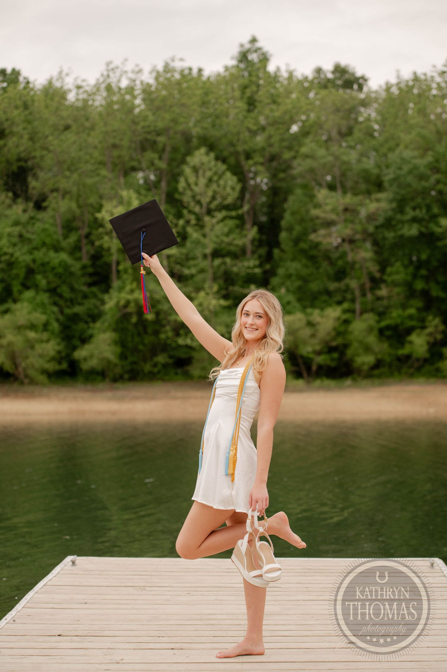 Graduate in white dress celebrates on wooden dock by lake holding cap with forest background.