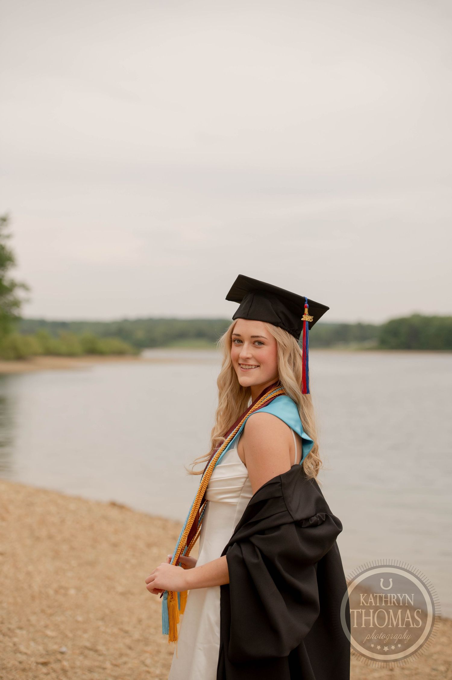 Graduate in cap and gown poses with honors cord by a lakeside during outdoor graduation photo session.
