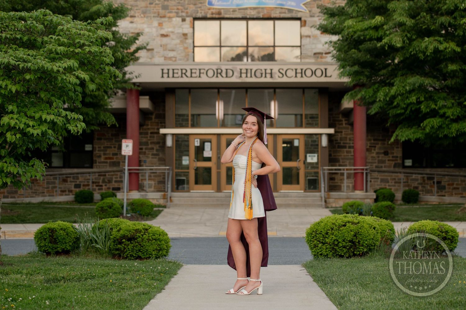 A graduate in white dress celebrates outside Hereford High School entrance while tossing graduation cap in the air.