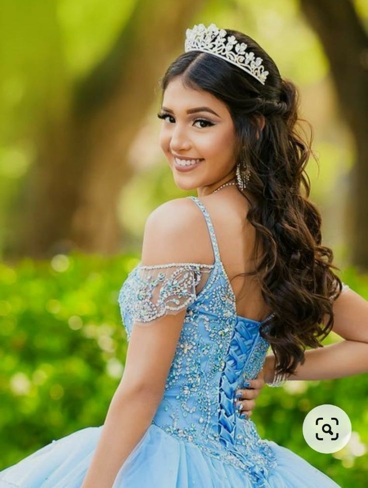 A quinceanera pose in a light blue embellished ball gown and crown against a green garden background.
