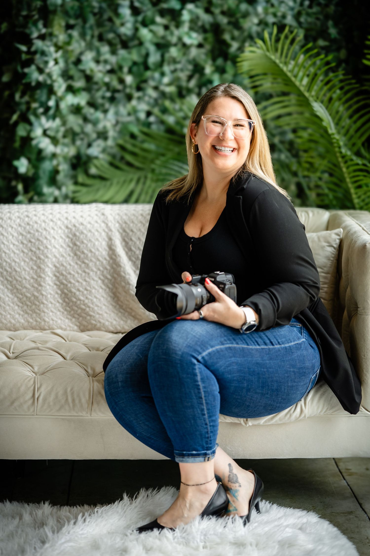 Professional photographer wearing black cardigan and jeans sits on white couch holding camera in indoor studio setting.