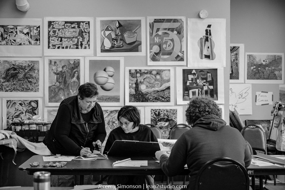 Students working together at a table in a classroom with various artistic prints and drawings displayed on the wall behind them.