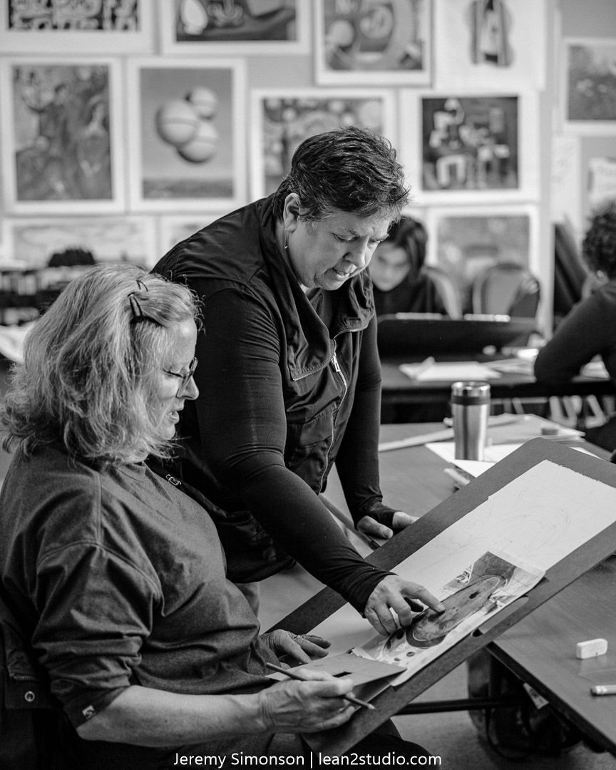 Two people collaborate over artwork at a desk in a studio setting with framed pieces displayed on the wall behind them.