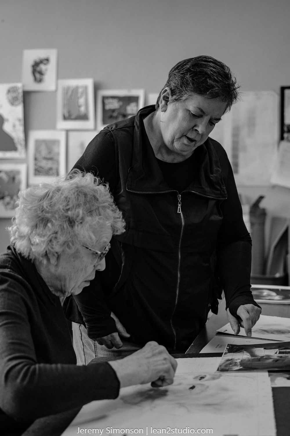 Two people examine artwork together at a desk in a black and white artistic workspace setting.