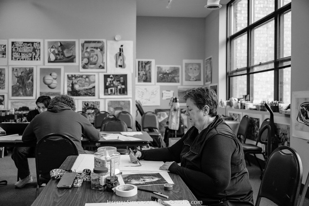 A black and white photo shows people working at desks in an office or study area with art displayed on the walls.