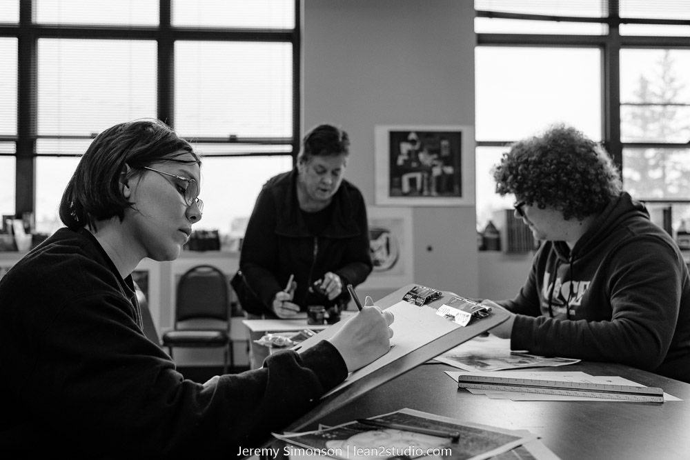Students work intently at their desks in a bright studio classroom with windows.