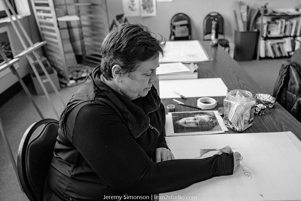 Person in black clothing works focused at desk with art materials and reference images.