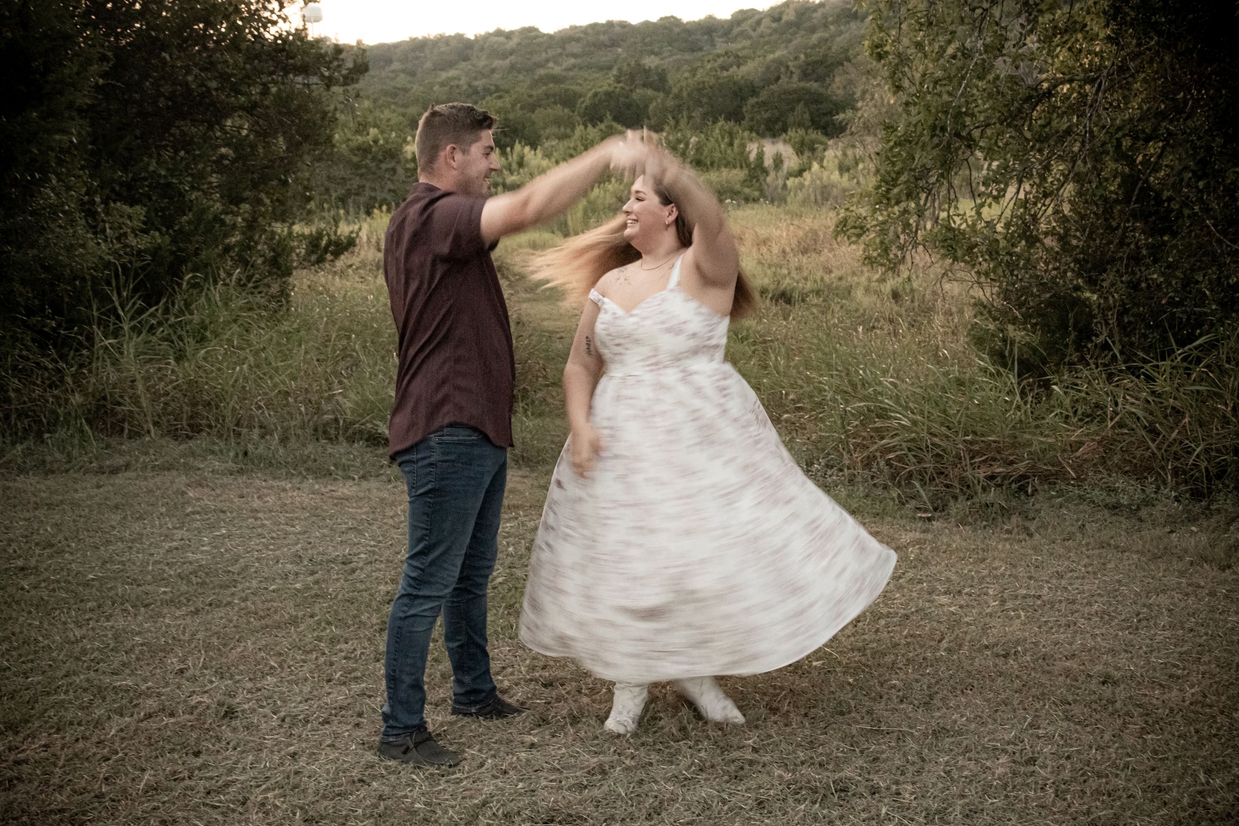 A couple dances outdoors in a field, woman in white dress spinning, surrounded by lush green trees at dusk.