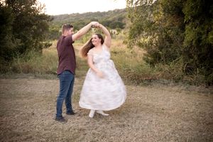 Couple dancing outdoors in a field, man spinning woman in white dress, surrounded by lush green trees at golden hour.
