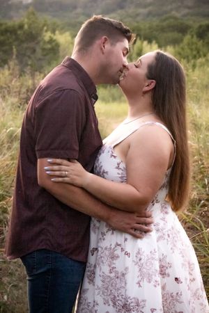 Couple sharing a romantic kiss outdoors in a green meadow, woman in floral dress, man in burgundy shirt.
