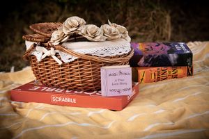 A wicker basket with paper flowers sits atop a Scrabble box and books on a yellow blanket outdoors.