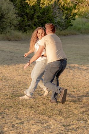 A smiling couple playfully chasing each other in a sunny field with green trees in the background.