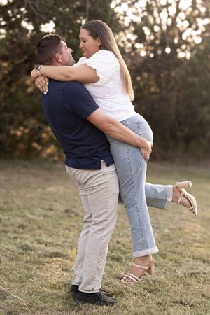 A man lifts a woman in an outdoor embrace, both smiling, surrounded by trees in a romantic couples photo.