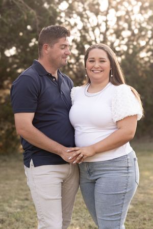 Smiling couple stands together outdoors in golden light, woman in white top and jeans, man in navy polo.