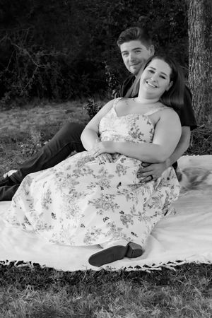 Black and white photo of a couple sitting on a blanket outdoors, woman in floral dress leaning against man near a tree.